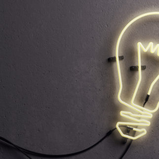 3D render of an light bulb shaped neon light against concrete wall. Neon light is turned on and it is glowing in lighter yellow color.